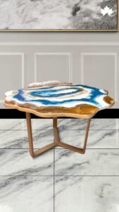 Luxurious handcrafted center table featuring an irregularly shaped top with swirling patterns of blue, white, and gold resin, resembling aerial views of a coastal landscape. The table is supported by a sleek bronze-colored metal base, adding a modern touch to the organically flowing design. This striking table combines functionality with artistic beauty, making it a standout piece in any contemporary interior.