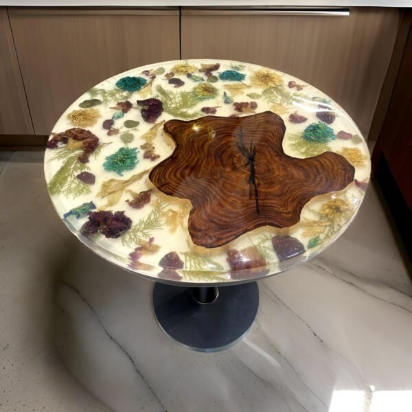 Unique round botanical inlay table featuring a stunning tabletop embedded with natural elements such as colorful dried flowers, seashells, and semi-precious stones, all set in a clear resin. At the center, a piece of beautifully grained wood adds a warm, organic touch. This table is supported by a sturdy dark grey pedestal base, making it a captivating centerpiece in a modern, light-filled space.