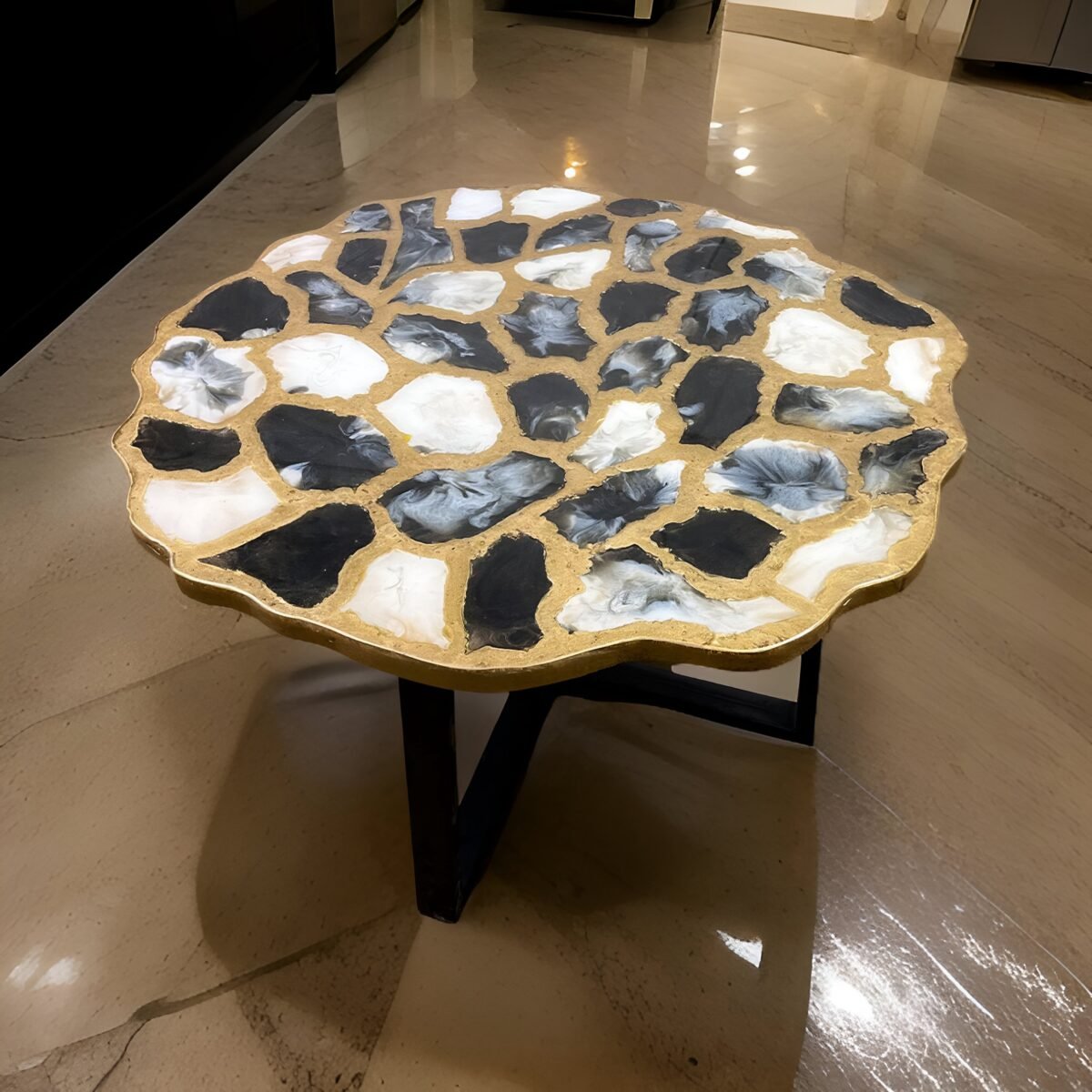 Artistic round coffee table featuring an eye-catching top designed with abstract black and white resin patches, surrounded by a golden trim. The table is supported by three sleek black legs, providing a modern contrast against the luxurious top. Set on a reflective marble floor, this unique table adds a bold visual element to any contemporary living space, blending artistic flair with functionality.