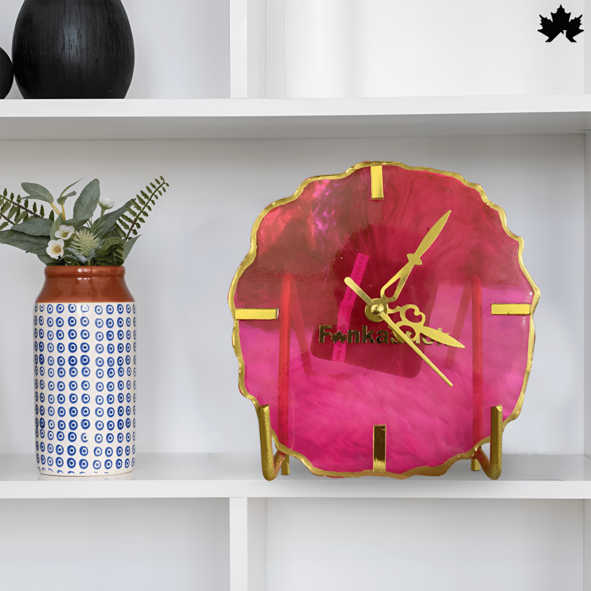 Elegant Fankaarlok designer clock made from vibrant pink resin, featuring gold accents and clock hands. The unique, irregular shape adds a modern touch to the home decor, displayed on a white shelf next to a blue and white patterned vase.