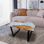 Contemporary center table featuring a unique design with a split surface; one half showcases natural wooden textures and the other half a flowing aqua blue resin that mimics water. The table is supported by sleek, black angular legs, perfectly complementing the modern and cozy living room setting with a plush taupe sectional sofa and a soft grey rug.