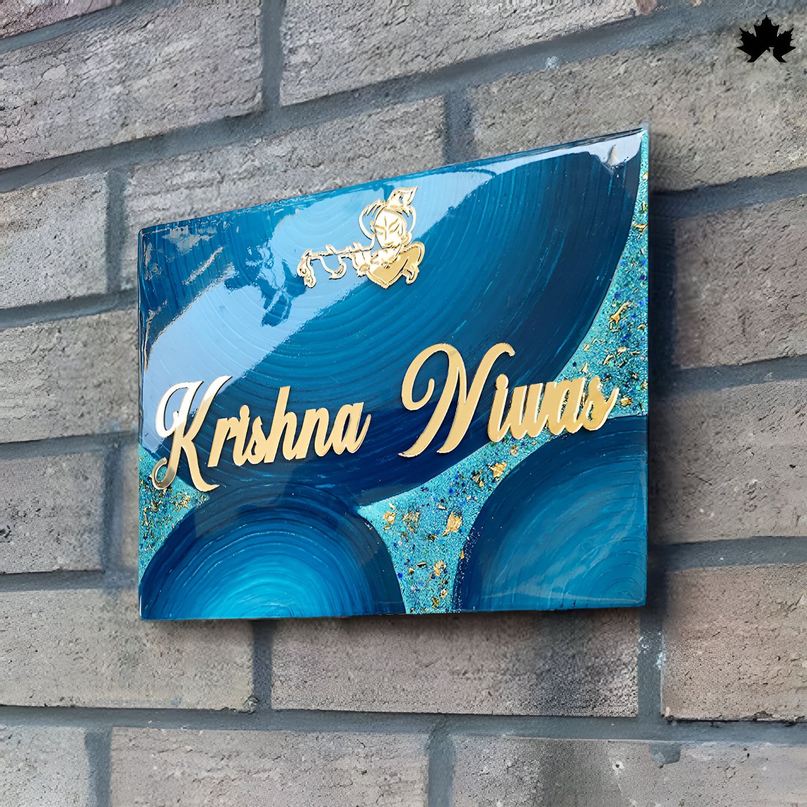 Custom door nameplates blue resin art wall plaque with gold leaf details featuring the name 'Krishna Nunu' in elegant script. The plaque shows swirling blue patterns with gold accents, mounted on a gray brick wall.