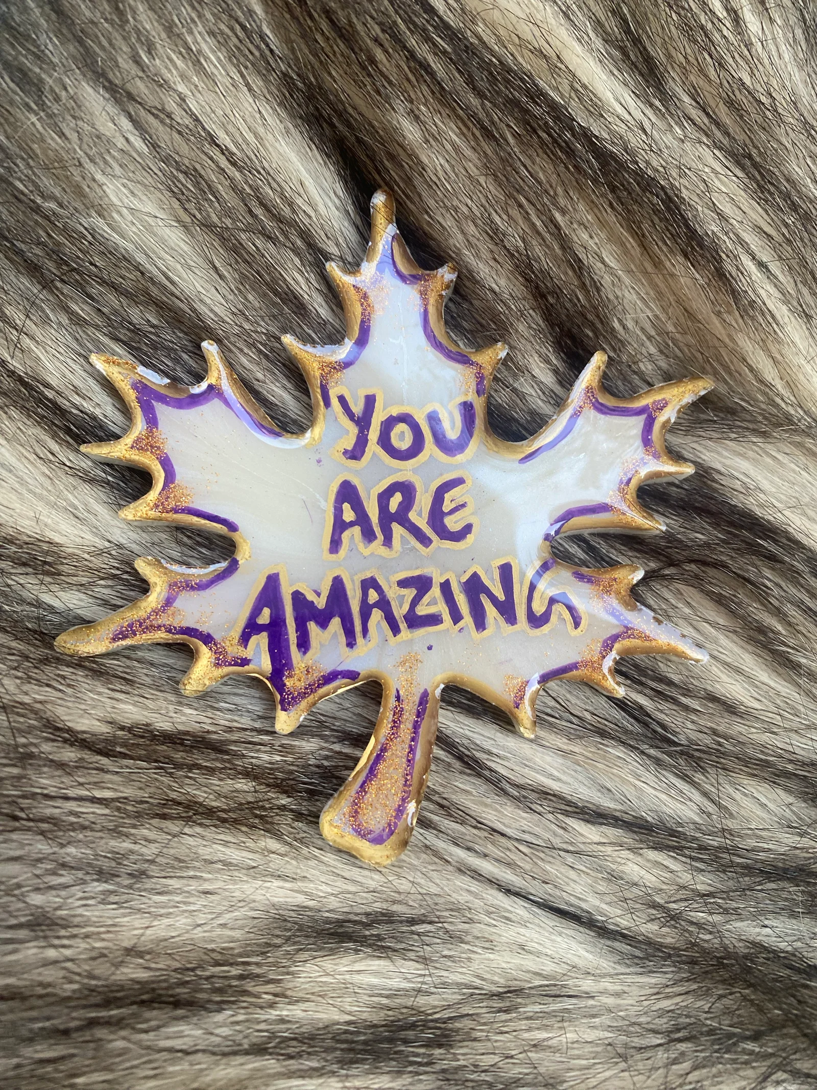 Custom-designed, leaf-shaped resin art piece with a motivational message reading 'You Are Amazing' in vibrant purple lettering, outlined with golden accents. The piece is showcased against a natural fur background, adding an organic touch to the inspirational and decorative item.