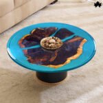 A stunning circular handcrafted resin & wood coffee table featuring a rich, wooden base with natural grain patterns. The tabletop is a vibrant blue resin that mimics a clear water body, encompassing scattered, wooden fragments, creating an illusion of islands. The resin's transparency enhances the depth and colors of the wood, making it a striking centerpiece in a cozy, carpeted living room.