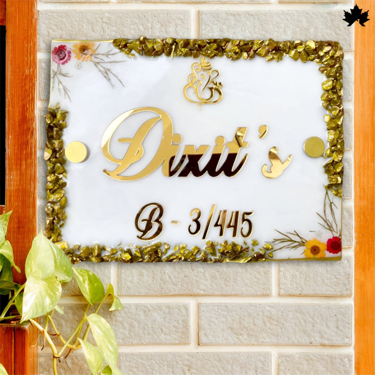 Elegant Custom Entrance Nameplate featuring the name in bold, cursive gold lettering, surrounded by a gold-edged, resin-filled frame adorned with small natural elements and floral motifs, mounted on a brick wall next to a lush plant.