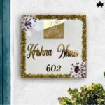 The image features the "Gold-Trim Luxury Villa Nameplate" from Fankaarlok, a stunning house nameplate. This piece elegantly displays the villa name "Krishna Niwas 602" in sophisticated golden lettering on a creamy white background, adorned with a delicately positioned golden image of Lord Krishna at the top left. The edges are beautifully embellished with a dense, glittery gold trim, adding a luxurious touch to the overall design, perfect for adding a regal look to any home entrance.