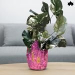 The image showcases a Blush Elegance Pink - Designer Plant Vase from Fankaarlok, featuring a textured pink resin body with gold leaf accents. This planter blends vibrant color and luxurious design, making it a striking piece for displaying plants and enhancing the decor of any modern living space.