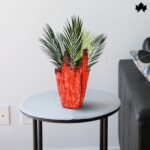 A vibrant and luxurious Scarlet Elegance Luxury Indoor Planter indoor planter from Fankaarlok, featuring a bold red resin body adorned with unique metallic accents. Perfectly positioned on a sleek table, this planter is filled with lush green palm fronds, enhancing the vivid contrast and adding a splash of sophisticated color to any modern living space.