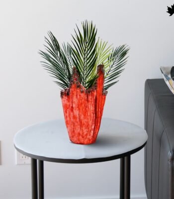 A vibrant and luxurious Scarlet Elegance Luxury Indoor Planter indoor planter from Fankaarlok, featuring a bold red resin body adorned with unique metallic accents. Perfectly positioned on a sleek table, this planter is filled with lush green palm fronds, enhancing the vivid contrast and adding a splash of sophisticated color to any modern living space.